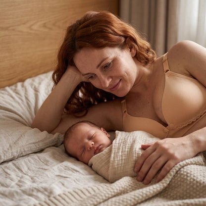 maman portant un soutien gorge d'allaitement allonger sur un lit a coter de son bébé endormi 
